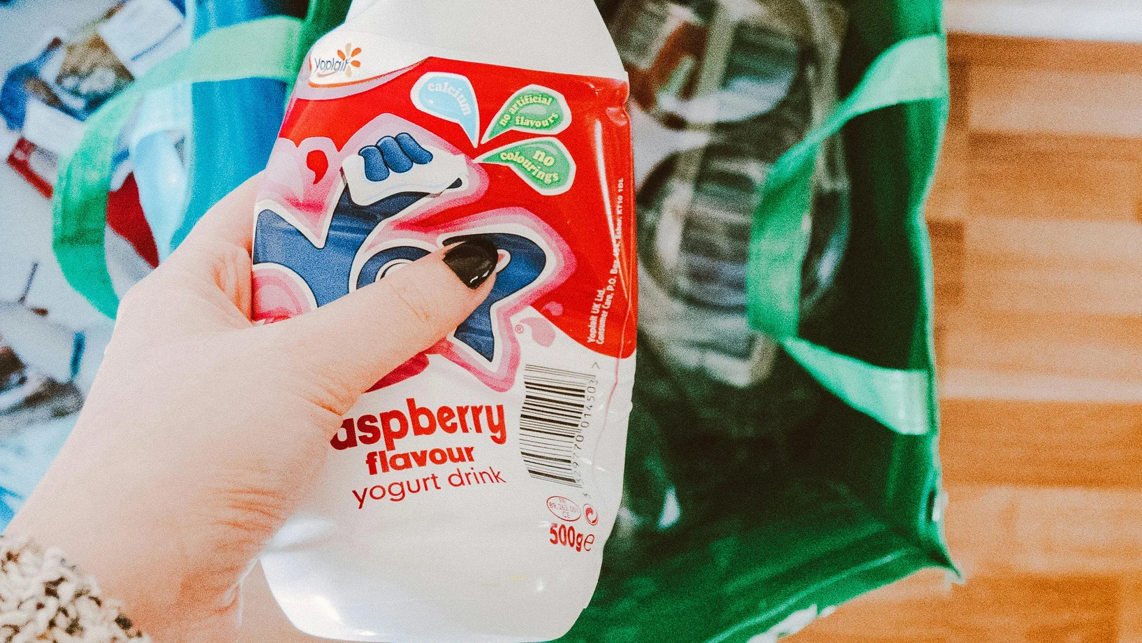 A person recycling a raspberry yogurt drink bottle into a green recycling bag.