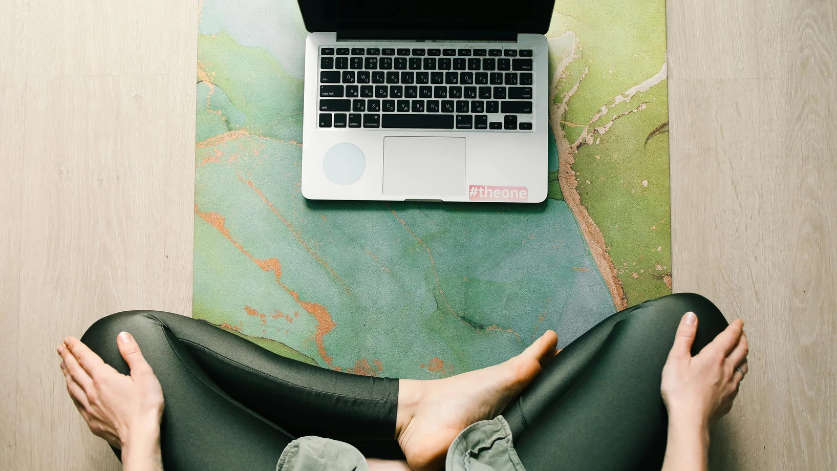 Adult sitting cross-legged on a yoga mat using a laptop, viewed from above.