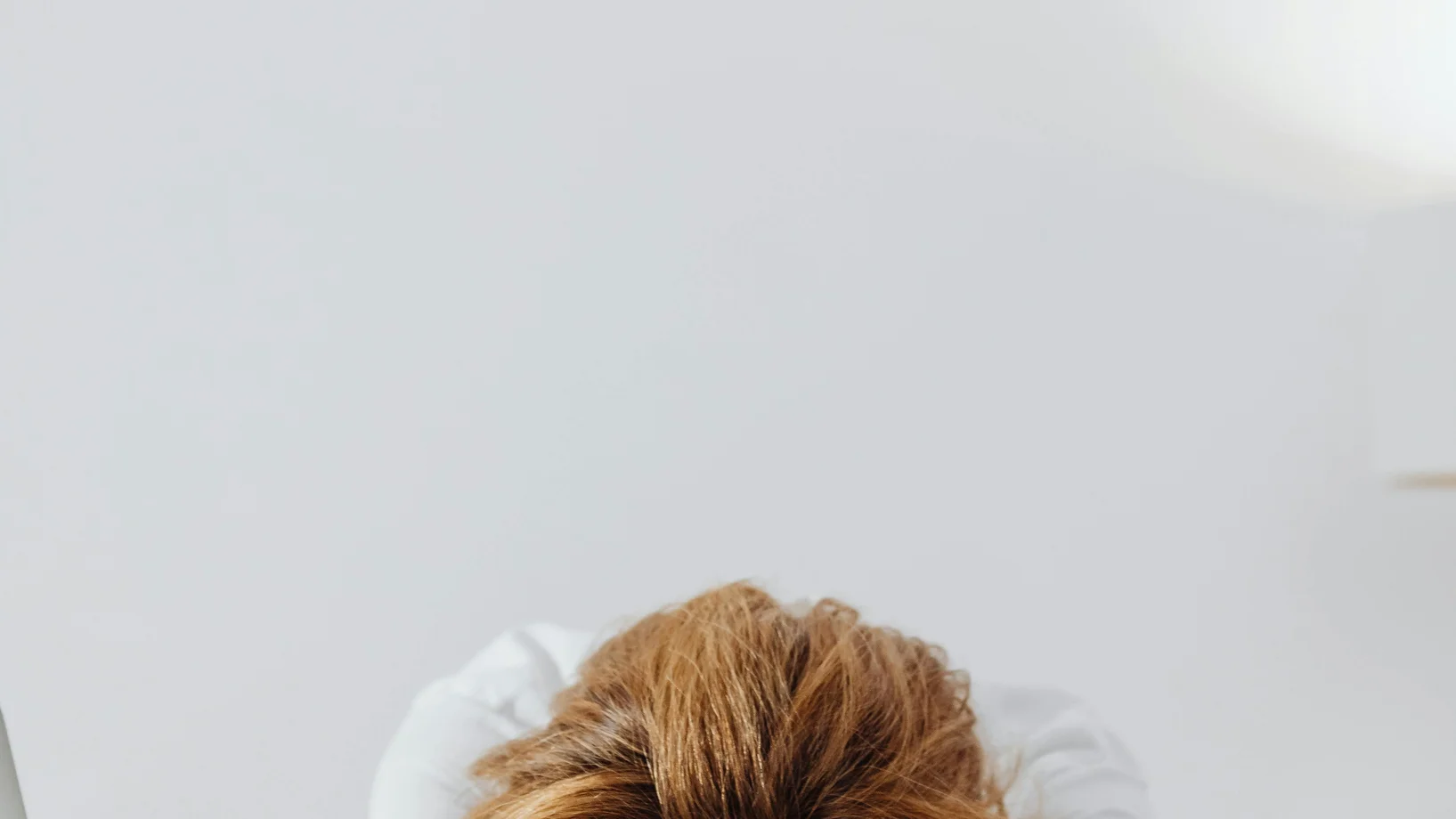 A tired woman with red nails resting her head on a white desk in a bright office setting.