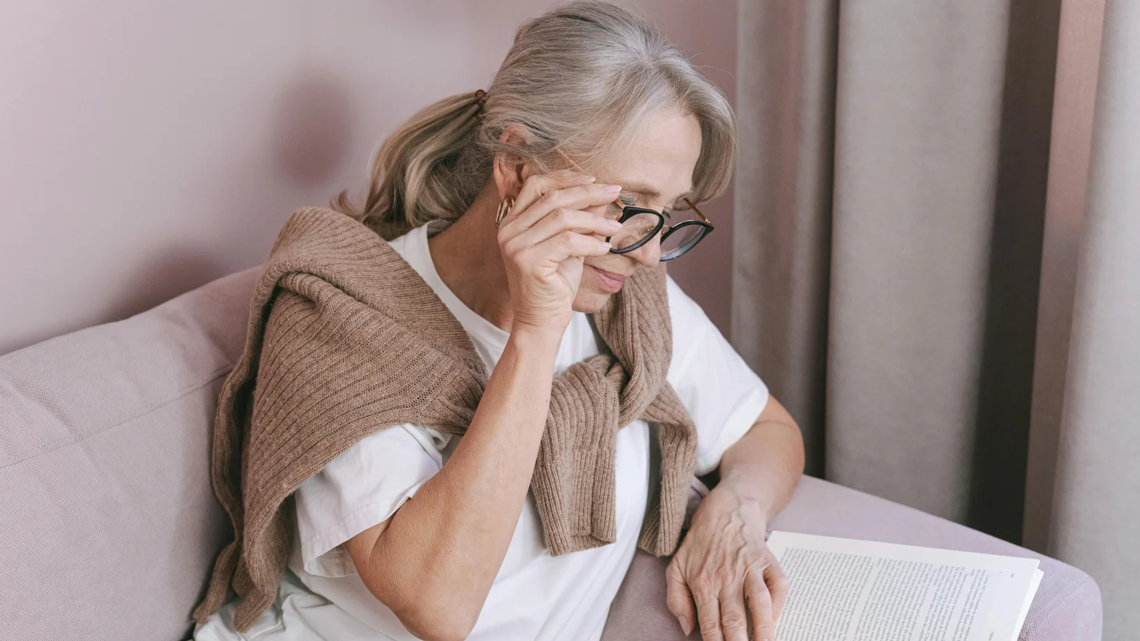 Elderly woman with glasses reading on a couch for leisure at home.
