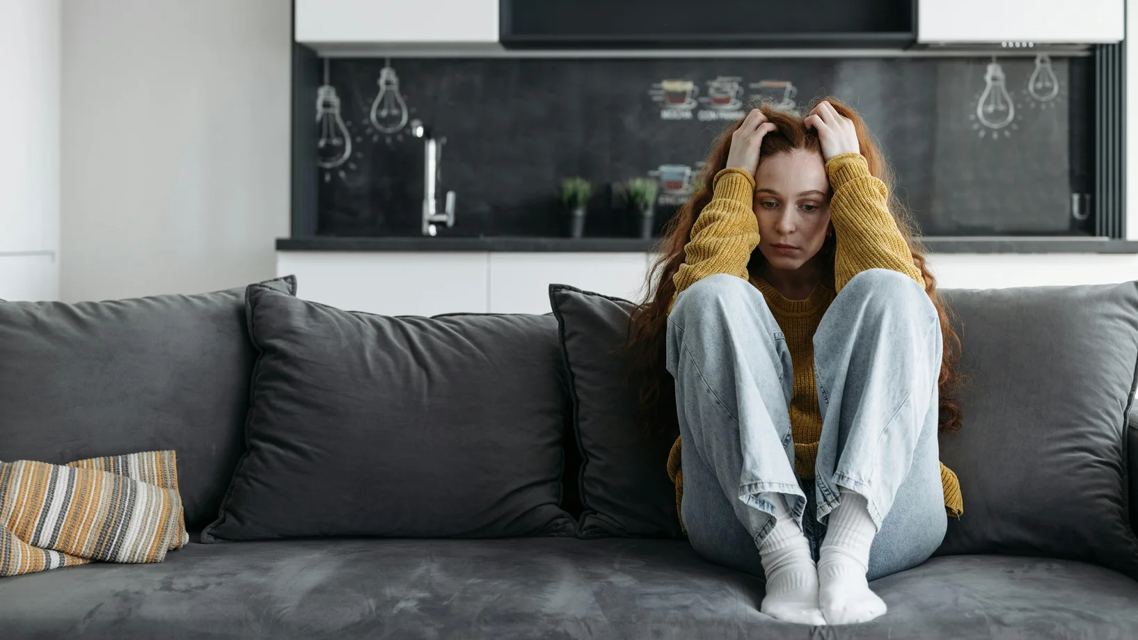 A woman in a yellow sweater sits on a couch, looking sad and pensive, indoors.