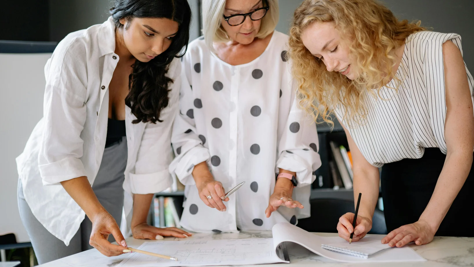 Three women working together over documents on a marble table indoors, showcasing teamwork and planning.