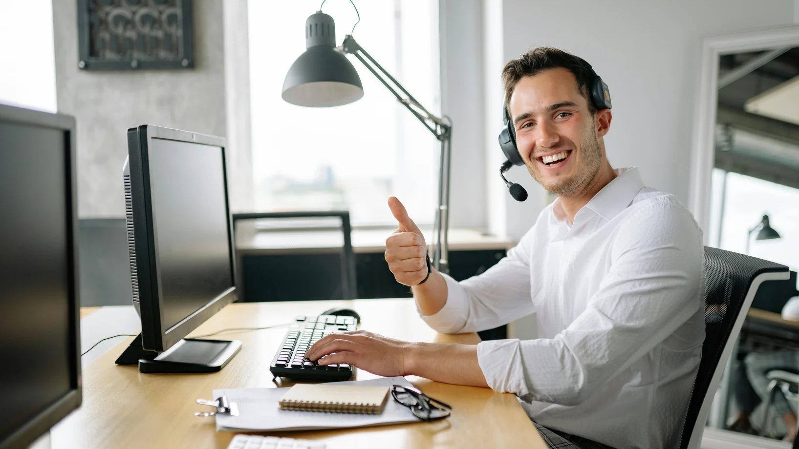 Happy man in office wearing headset, giving a thumbs up while typing at a computer.