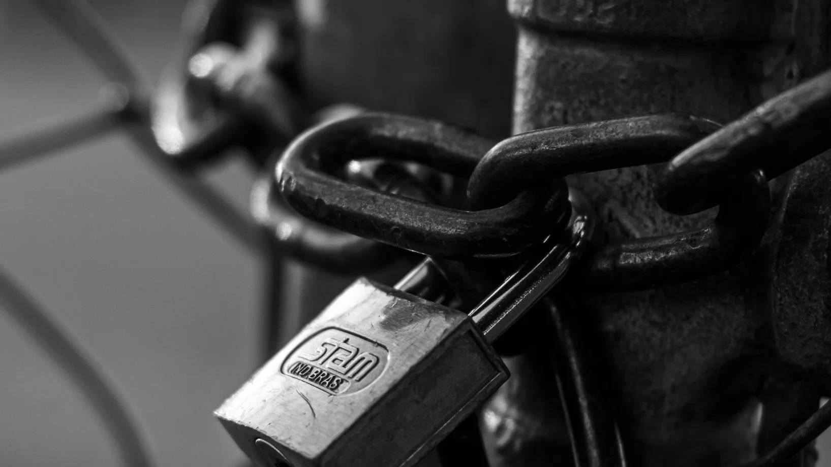 A detailed black and white image of a metal padlock securing a gate chain.